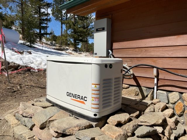 Generac generator, white and beige, installed next to a wood-sided building on a rocky foundation. Snow in the background.
