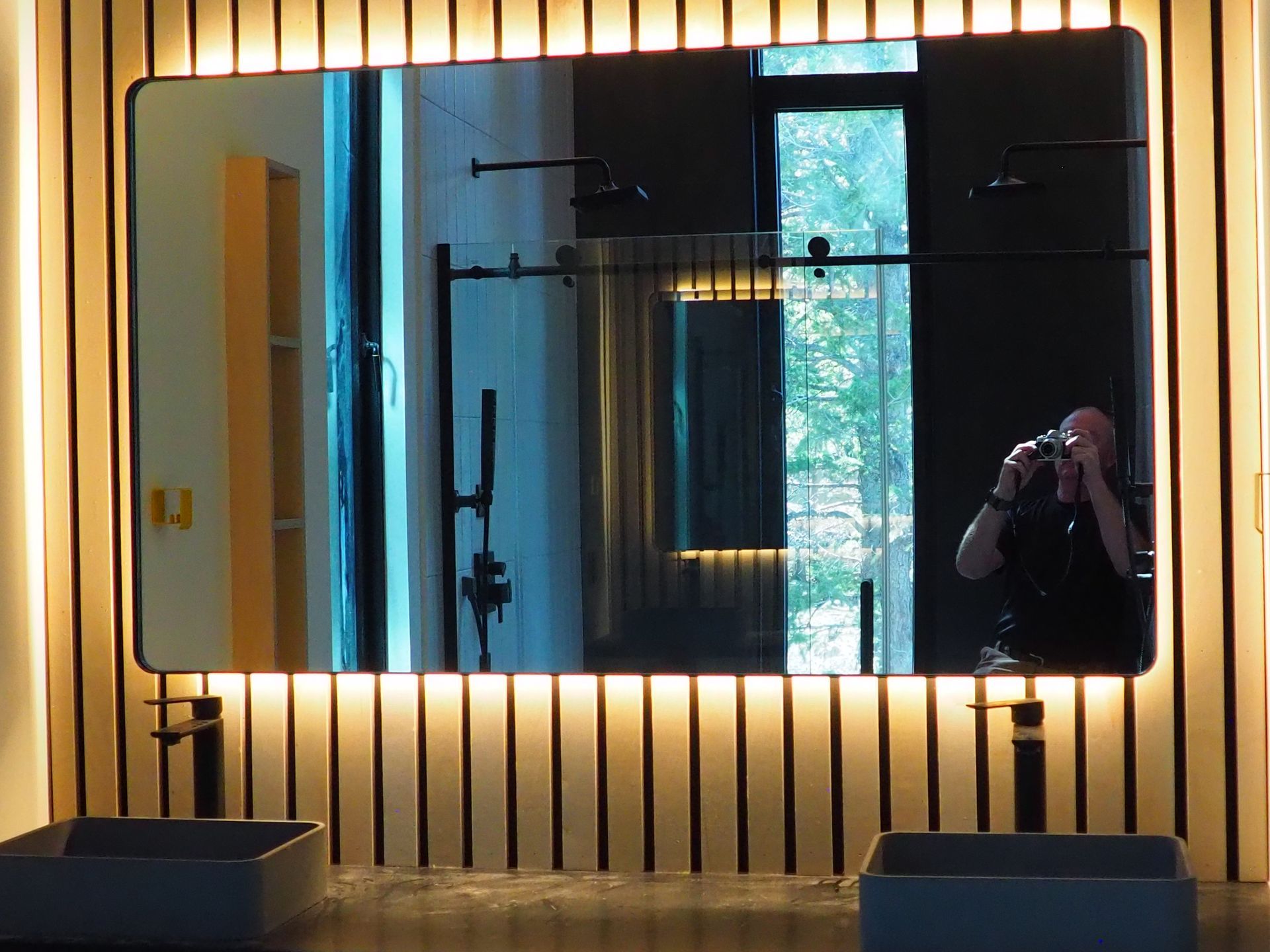 Bathroom mirror with person taking photo, reflected in mirror above two sinks. Yellow cabinet and black fixtures.