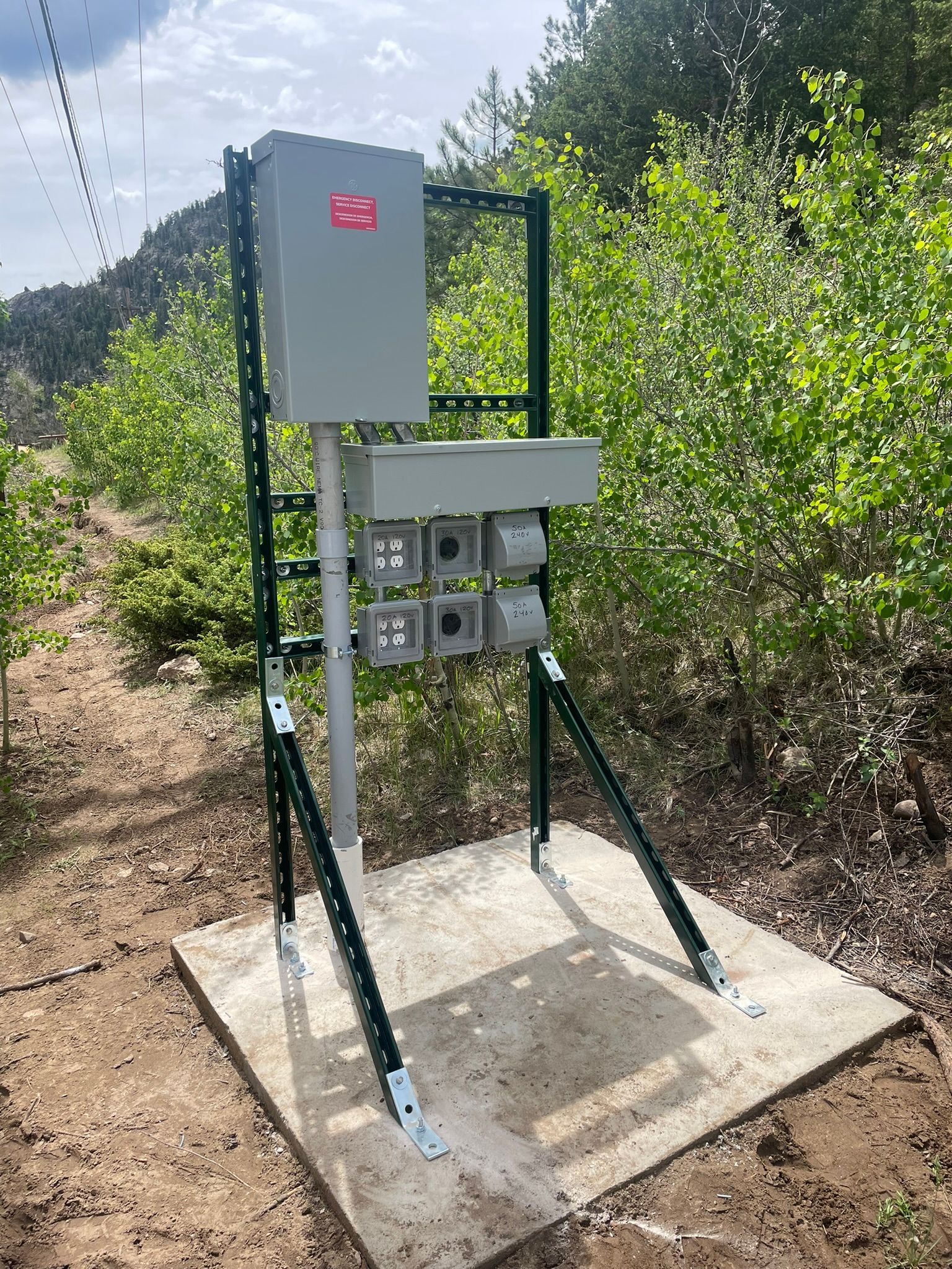 Electrical equipment box on a green metal frame, mounted on a concrete pad, outdoors with trees.