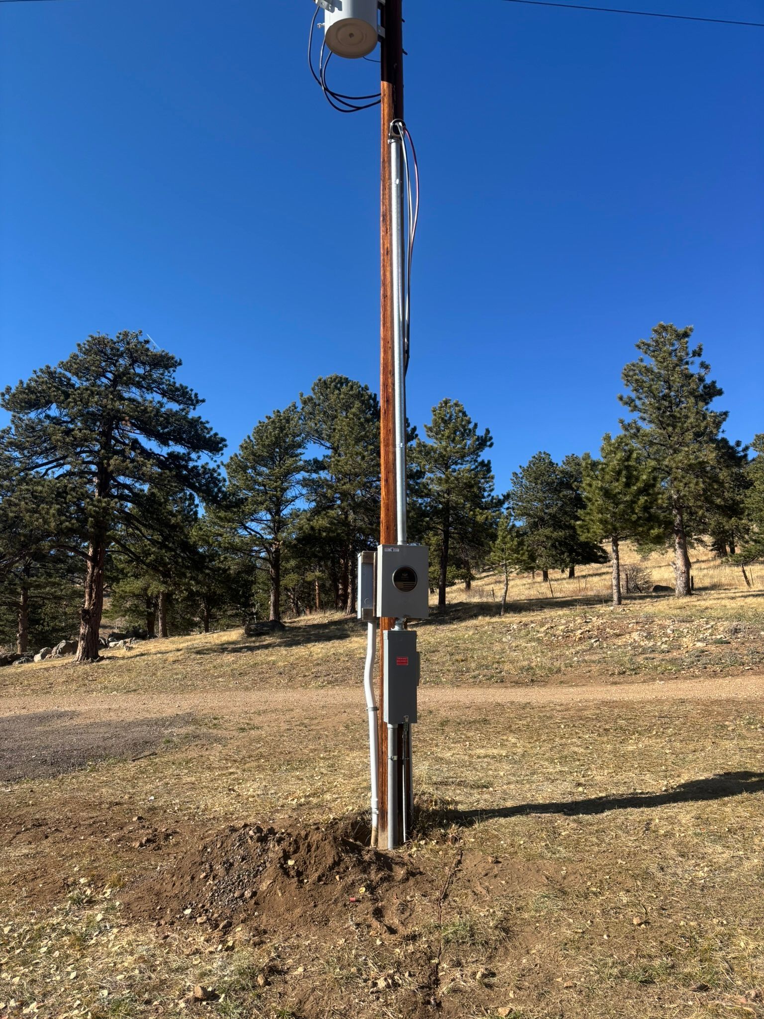 Utility pole with electrical boxes, antennas, set in a grassy area with trees under a blue sky.