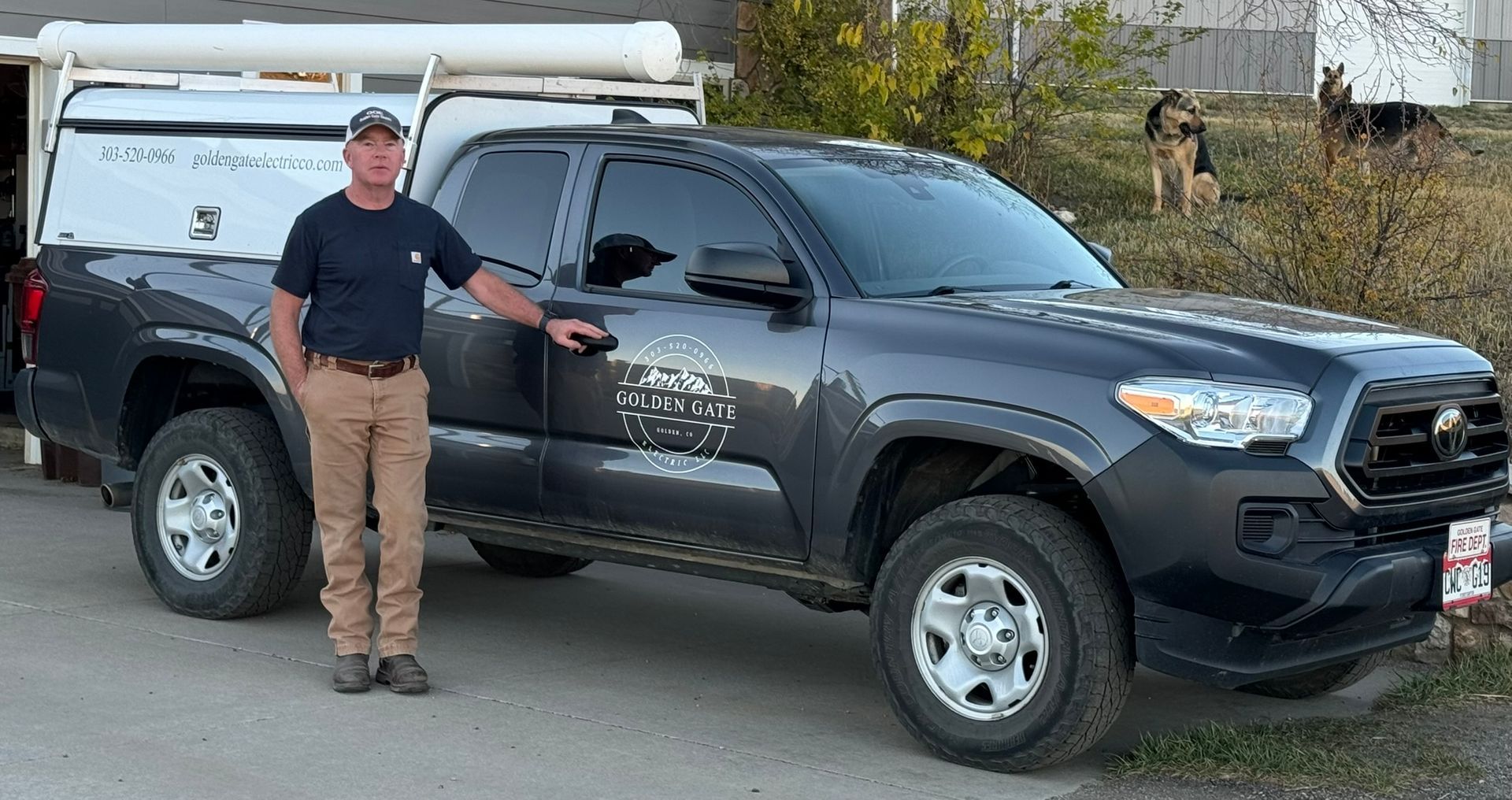 Man standing next to a dark gray pickup truck with a white truck bed cover, a dog is in the background.