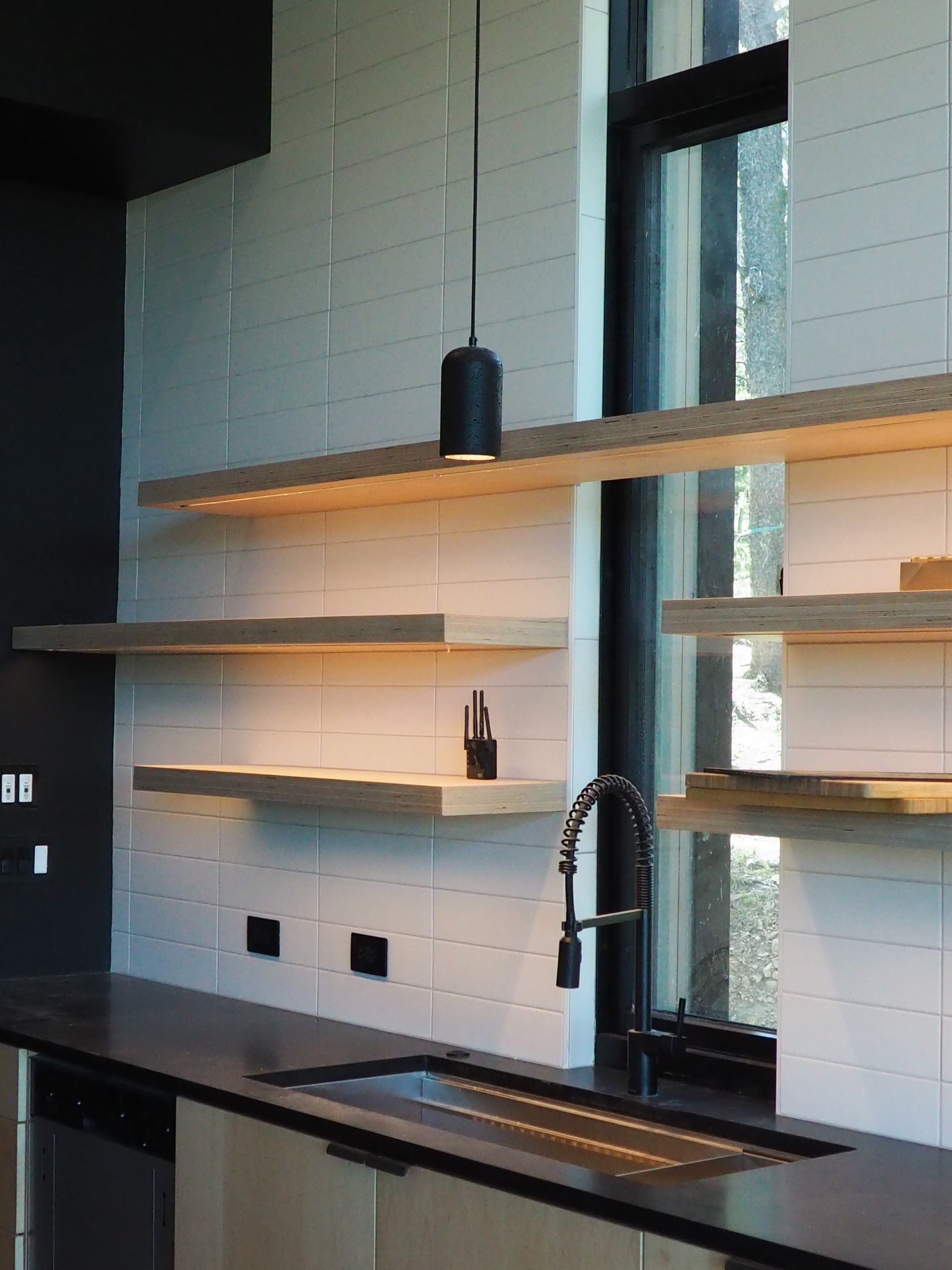 Modern kitchen with floating shelves, black countertop, and sink by a window.