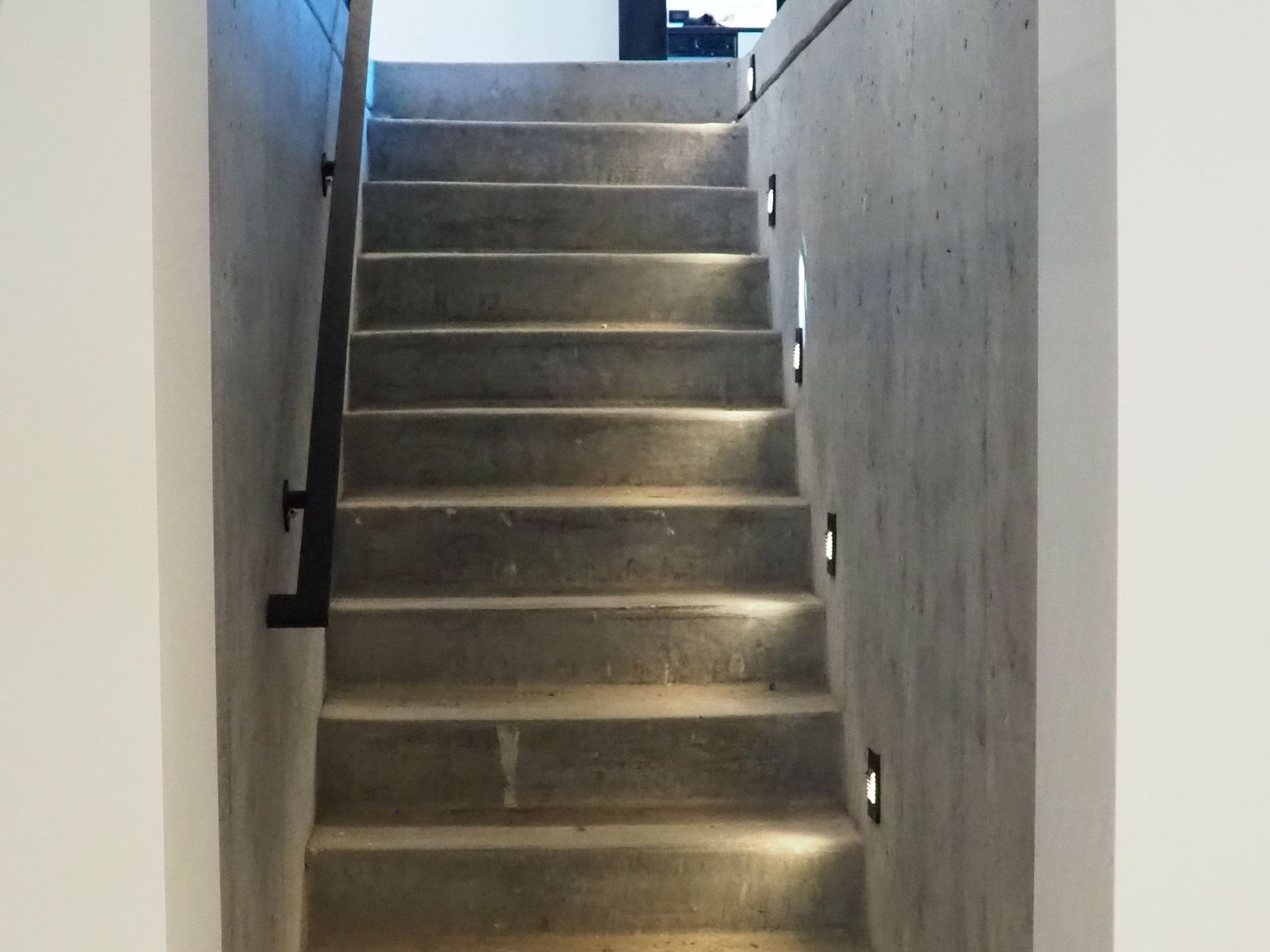 Concrete staircase with recessed lighting along the side wall, leading upwards. Black handrail on the left.