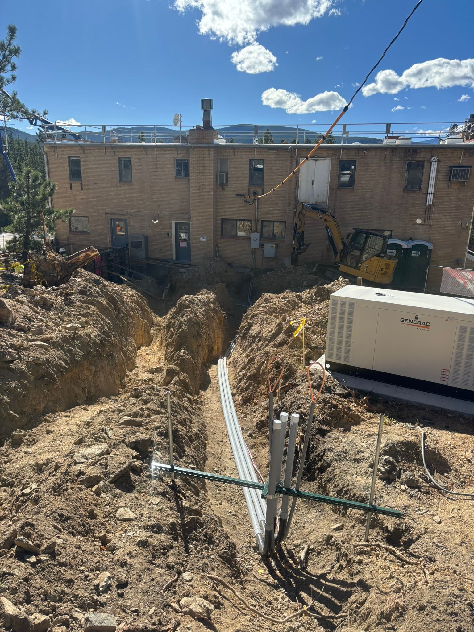 Construction site: Trench dug near a brick building. Conduit and equipment visible. Sunny day, blue sky.