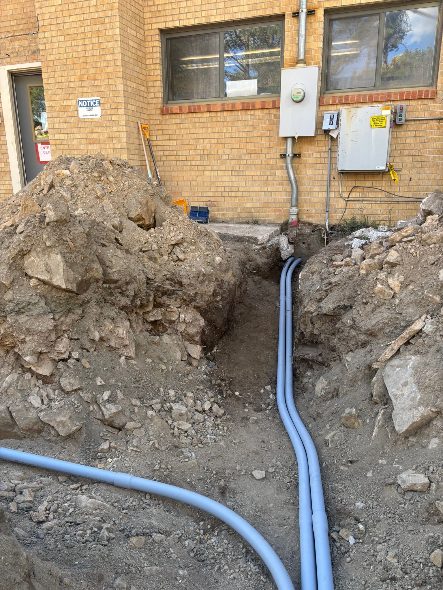 Trench dug next to a brick building; two blue conduits run from electrical boxes to the trench.