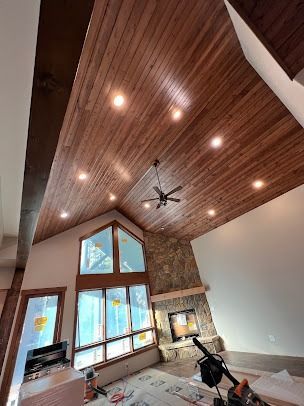 Wooden ceiling with recessed lights, stone fireplace, large windows in a room.