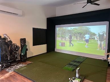 Golf simulator room with screen displaying golf game; green turf, golf bags, and hitting mat.