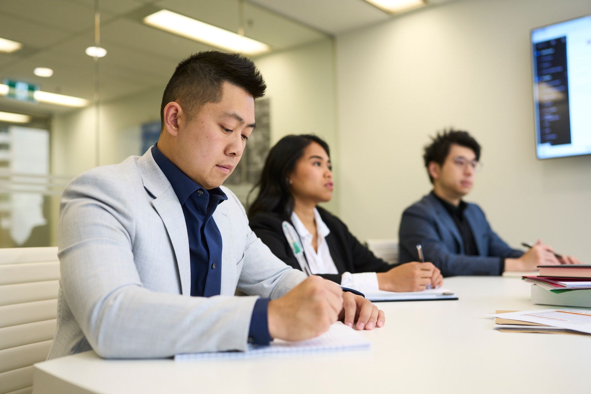 Three professionals in business attire sit at a white table in a bright office, focused on taking notes during a meeting.