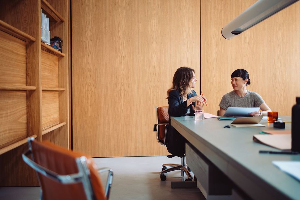 Two people sitting across a large conference table in a modern, wood-paneled office, engaged in a discussion