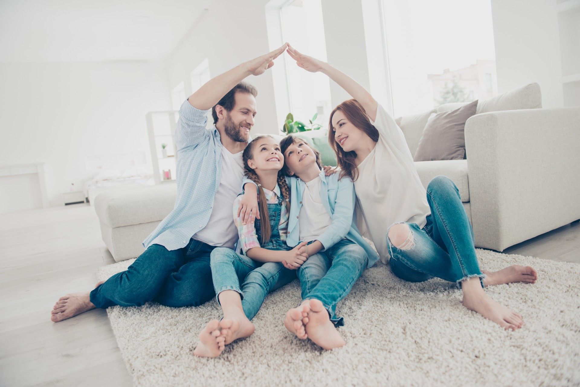 A family is sitting on the floor in a living room making a roof with their hands.