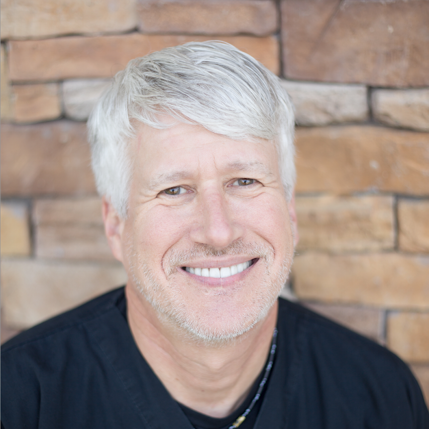 A man with gray hair is smiling in front of a brick wall.