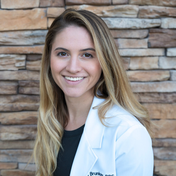 A woman in a lab coat is smiling in front of a brick wall