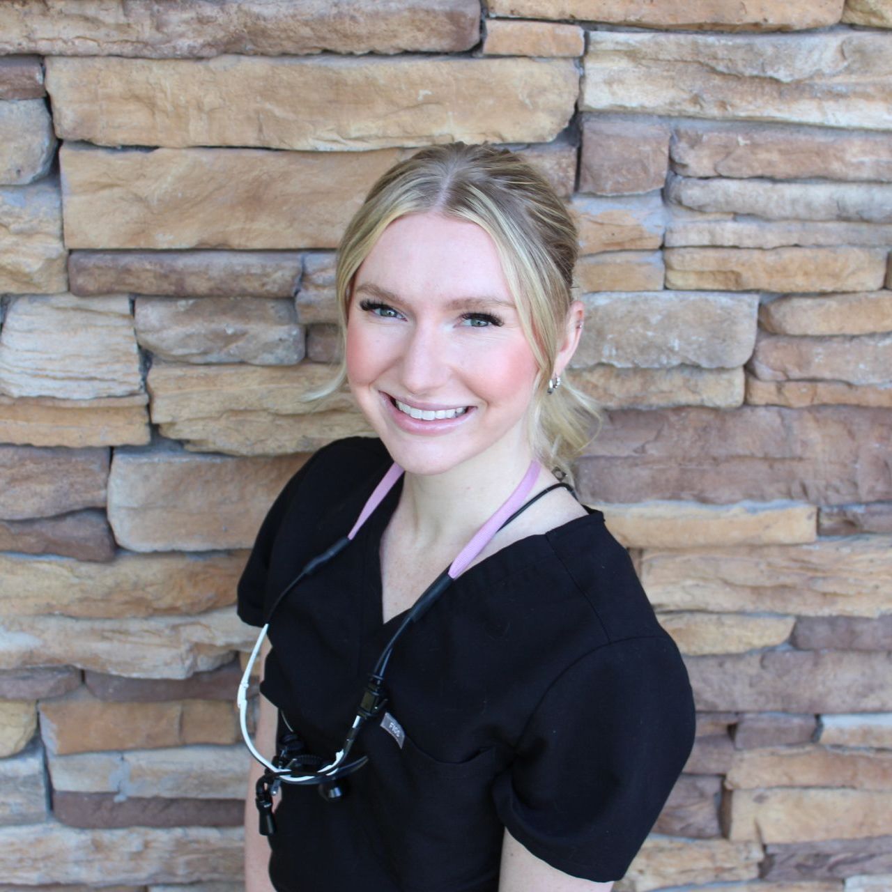 A woman with blonde hair and blue eyes is smiling in front of a brick wall.