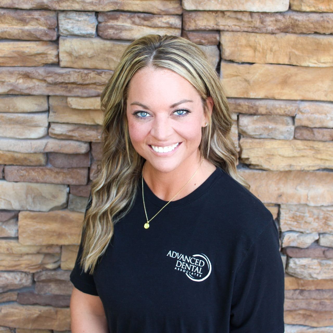 A woman with blonde hair and blue eyes is smiling in front of a brick wall.