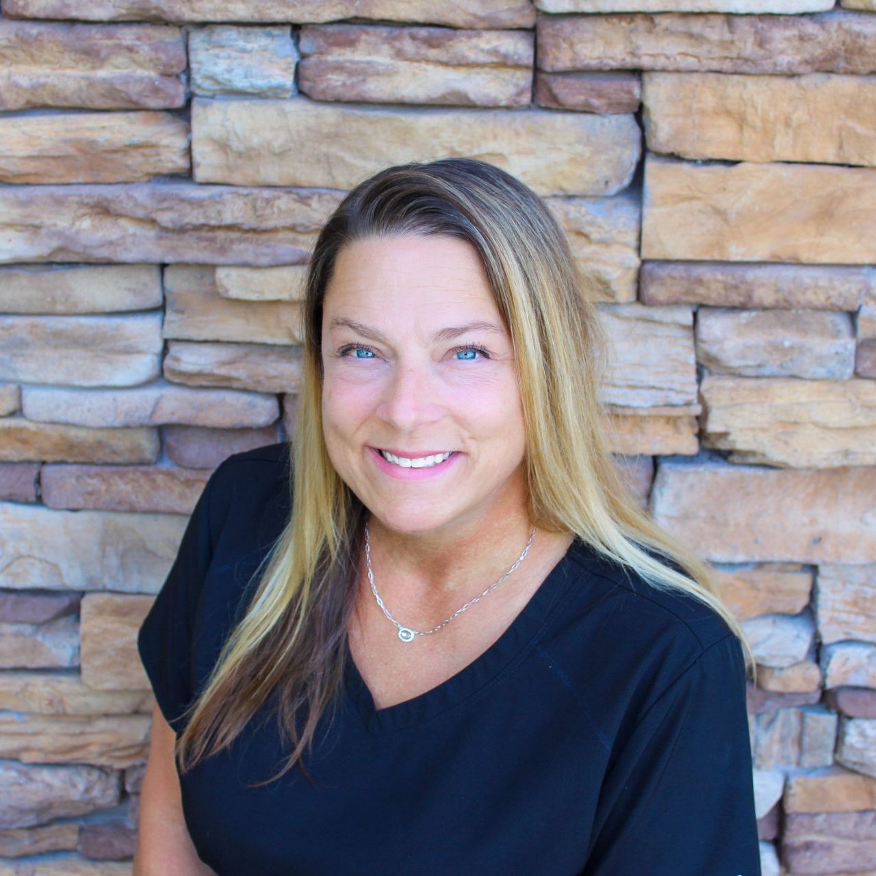 A woman in a black shirt is smiling in front of a stone wall.