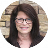 A woman wearing glasses is smiling in front of a brick wall.