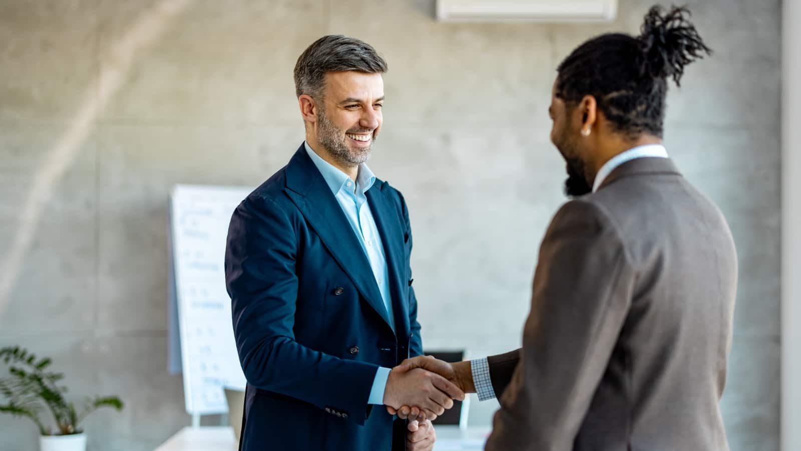 Two men in suits are shaking hands in an office.