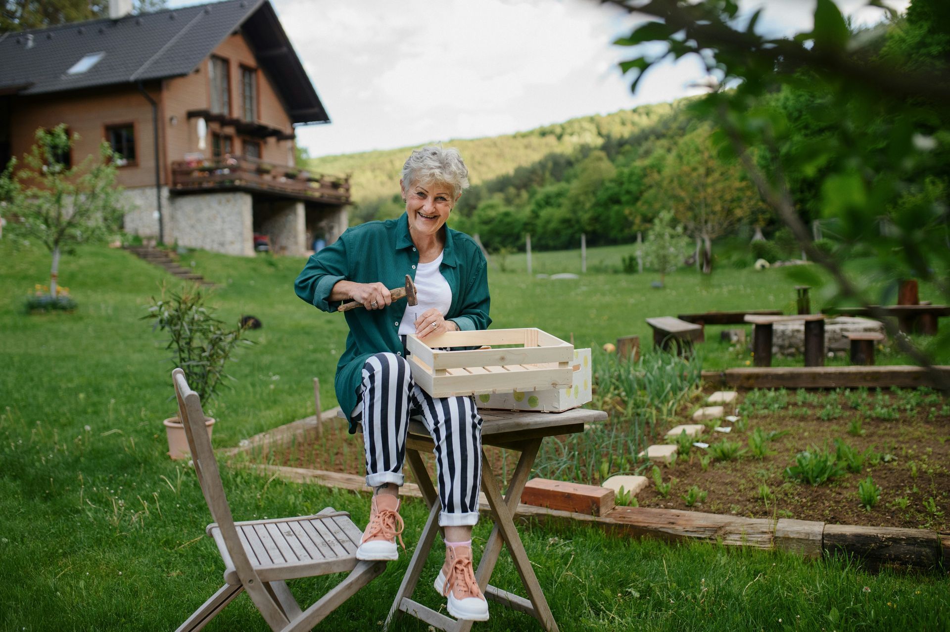 An elderly woman is sitting at a table in a garden in front of a house.