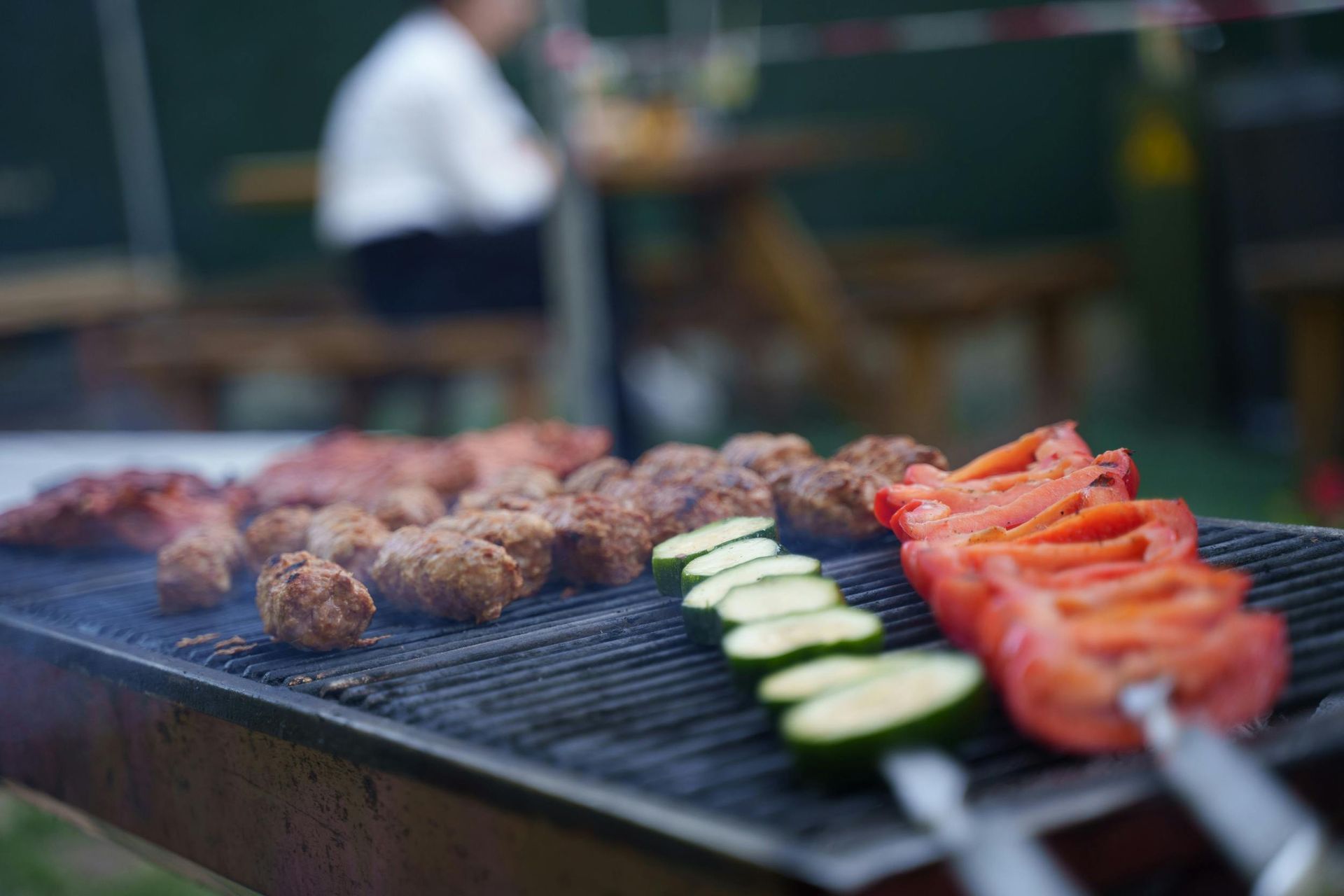 Grilled meat, zucchini, and tomato skewers sizzling on a barbecue with a blurred chef in the background