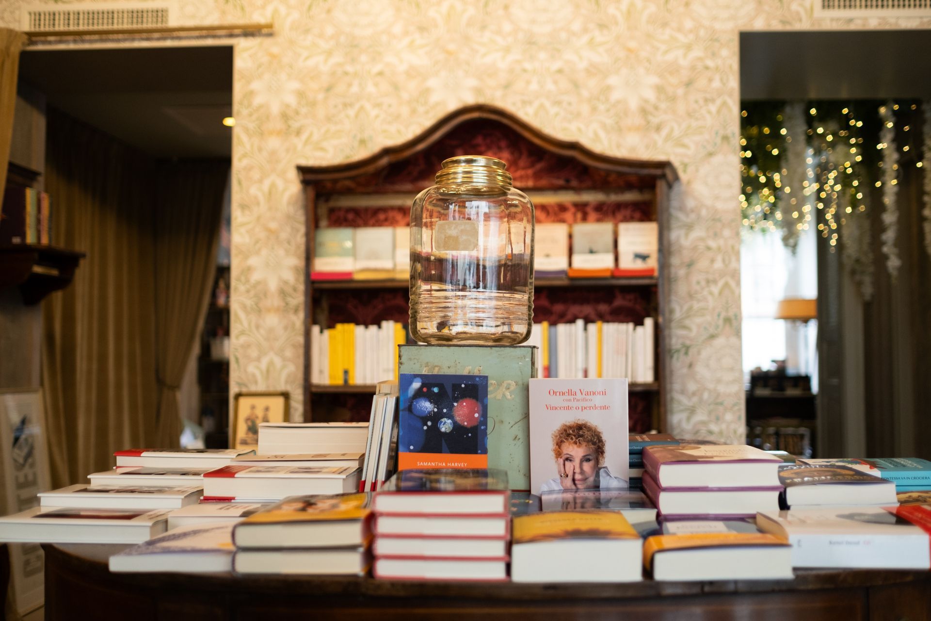 Una composizione di libri su un tavolo con pile di volumi e una lampada a sospensione accesa in una stanza accogliente.