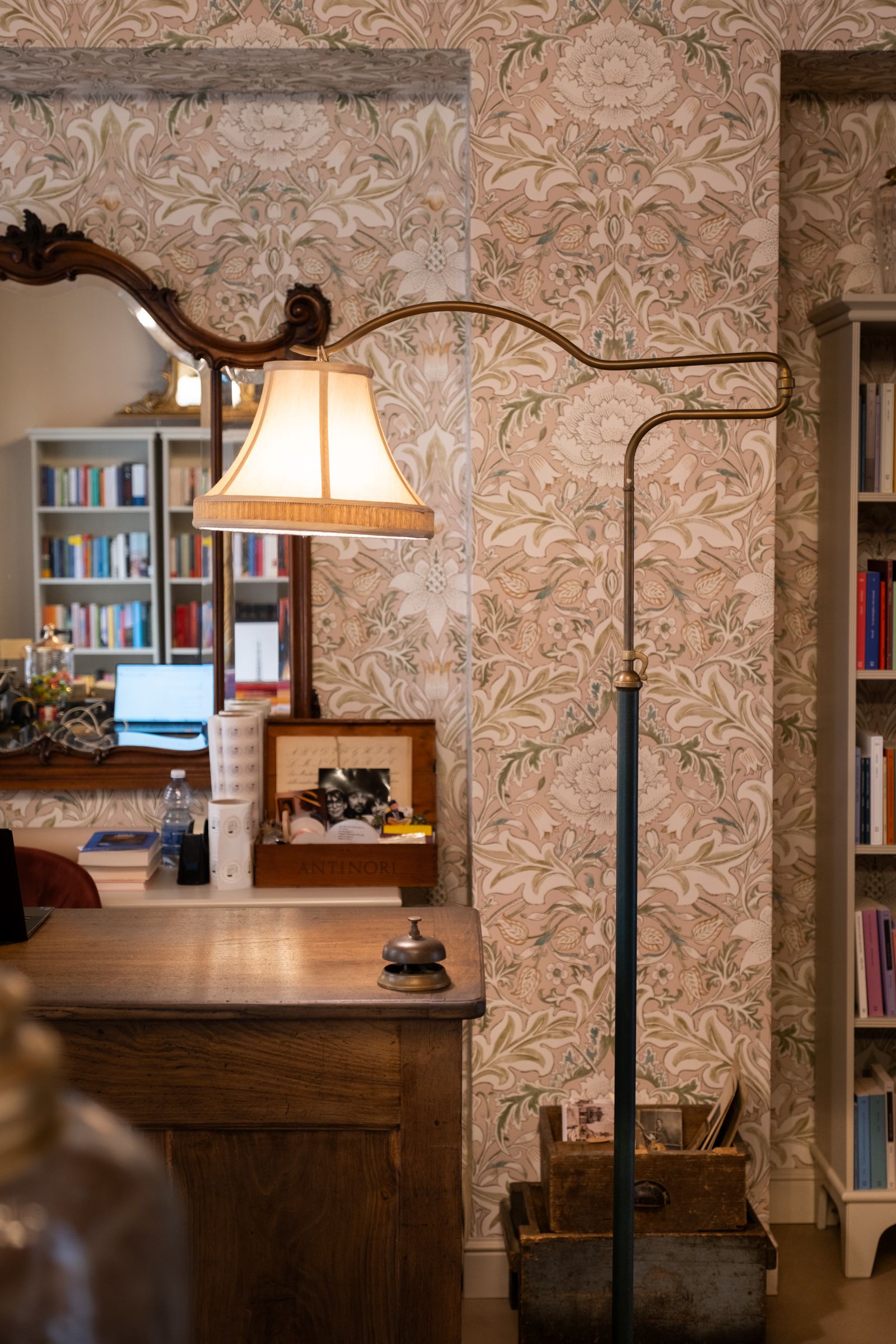 Interno accogliente con lampada a sospensione, tavolo in legno, carta da parati floreale e mensole visibili attraverso un arco.