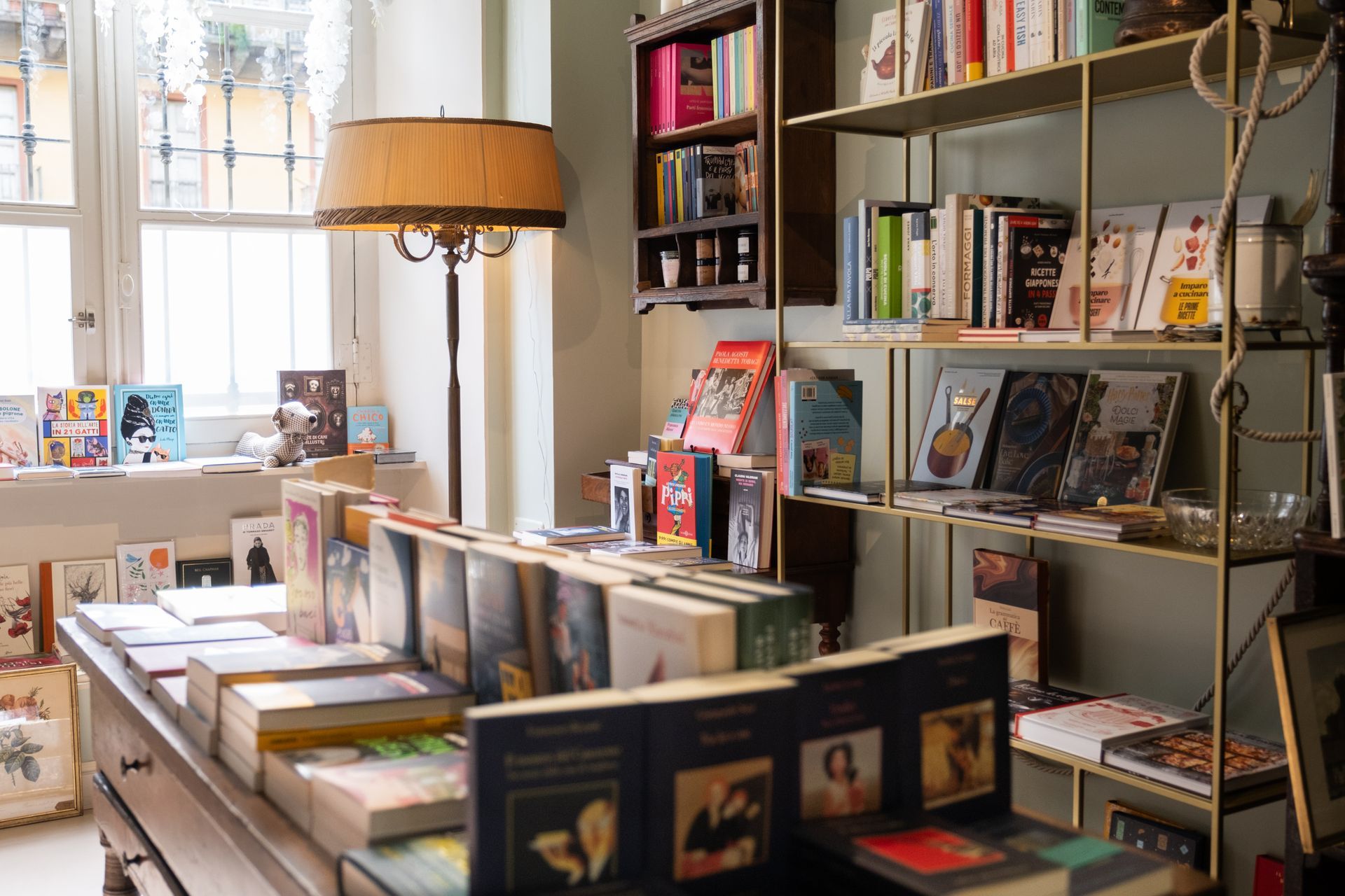 Libreria con scaffali pieni di libri, un tavolo espositivo e una lampada da terra dalla luce calda accanto a una finestra illuminata dal sole.