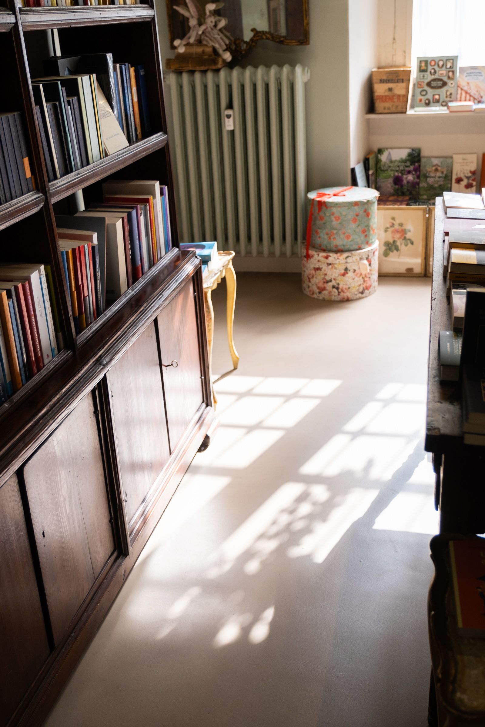 Stanza luminosa con librerie, termosifone, pouf decorato e ombre luminose proiettate dalle finestre sul pavimento.