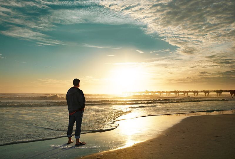 A man is standing on the beach looking at the sunset.