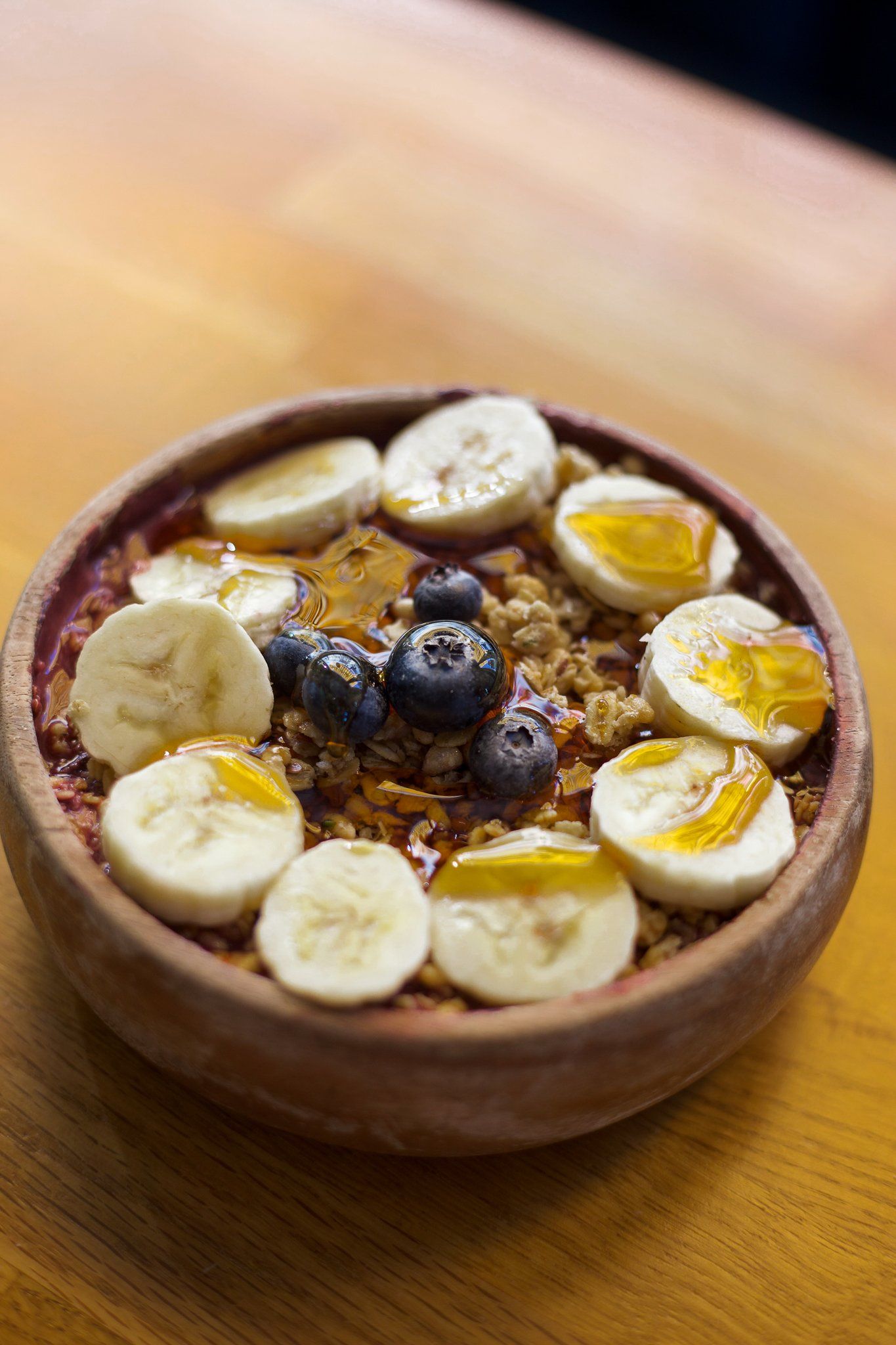 A bowl of food with bananas , blueberries , granola and honey on a wooden table.