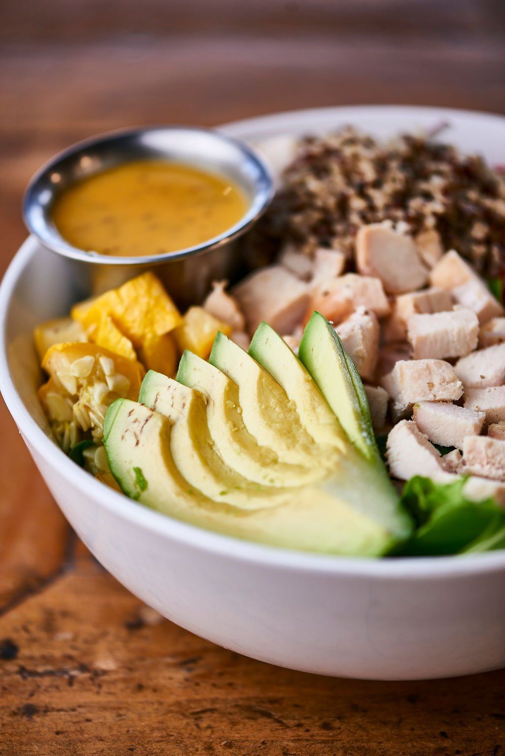 A bowl of food with avocado , chicken , rice and a dipping sauce on a wooden table.