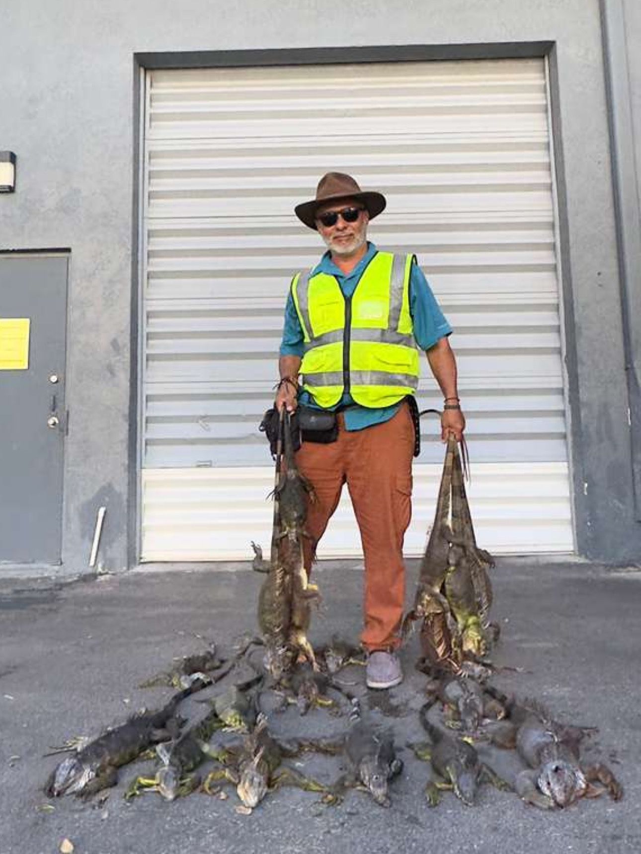 Man holds several dead iguanas, standing in front of a building with a roll-up door.