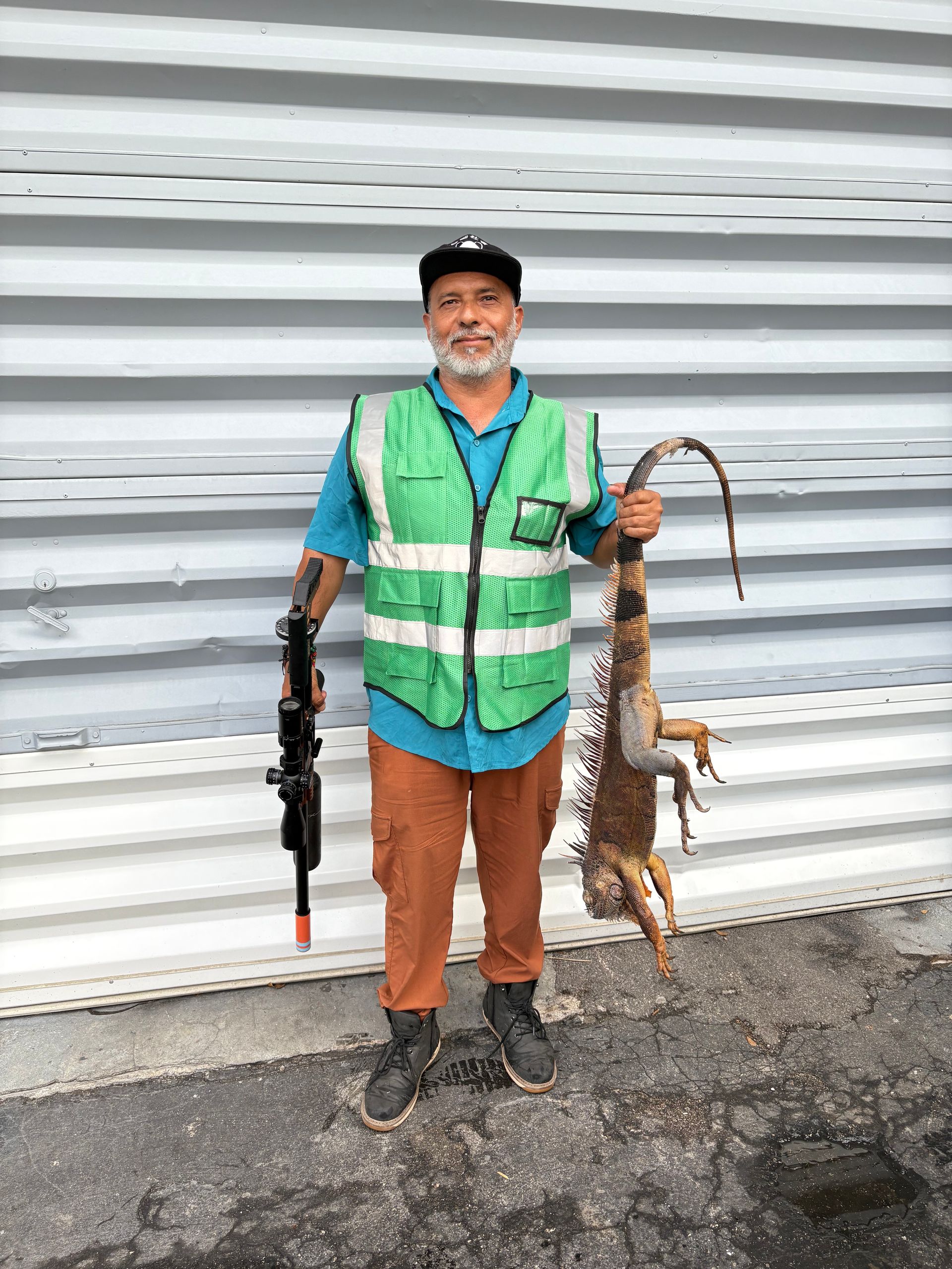 Man in safety vest holds a gun and a large iguana in front of a corrugated wall.