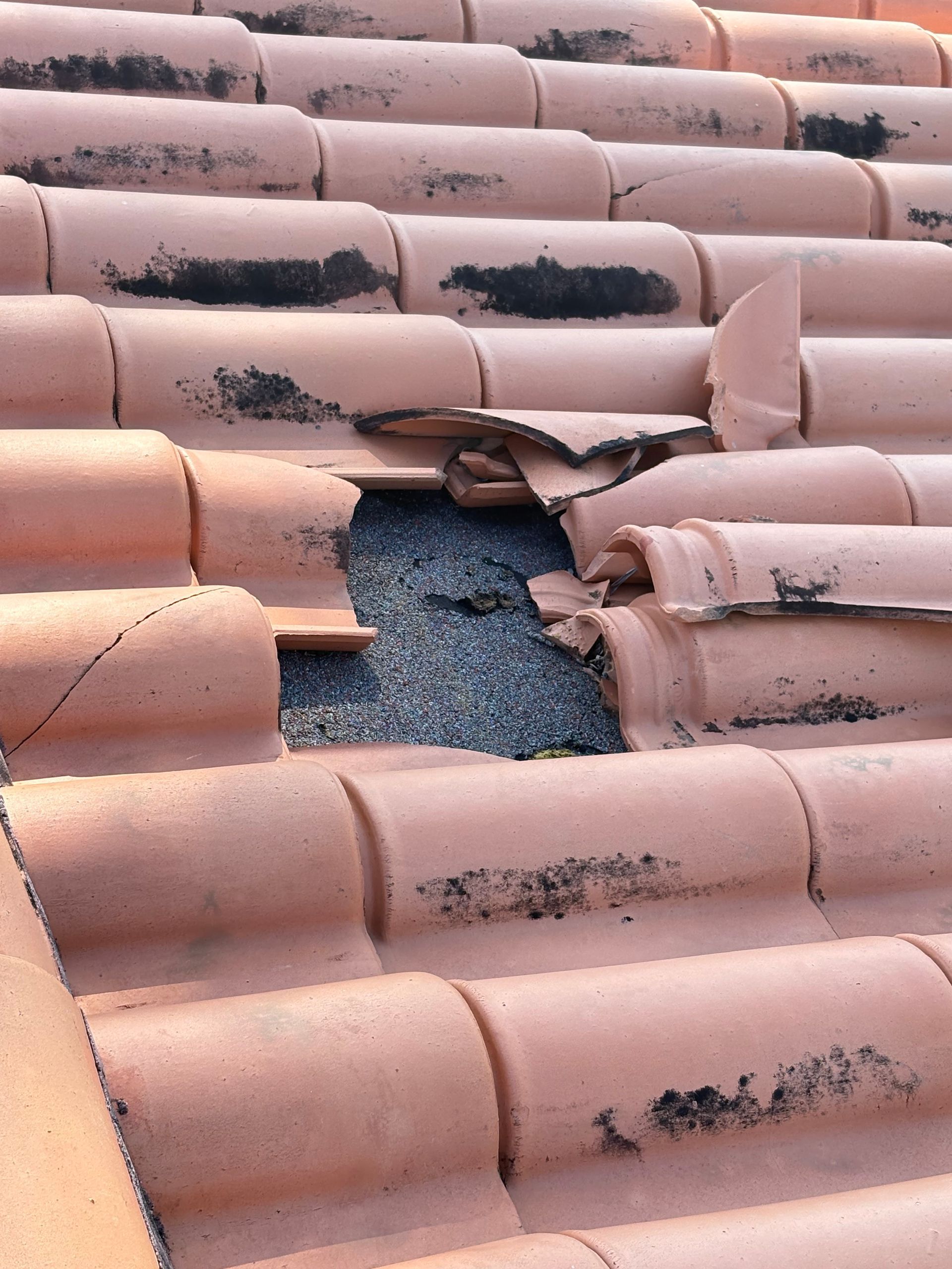 Damaged terracotta roof tiles with missing pieces exposing underlying gravel.