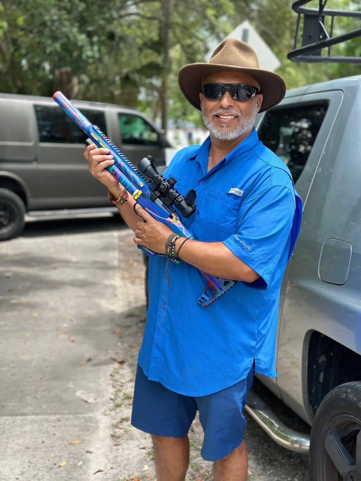 Man in blue shirt and hat holds a blue and purple rifle with a scope, standing by a truck.