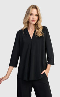 Woman in a black V-neck top and pants. She has long, wavy blonde hair and is standing against a light background.