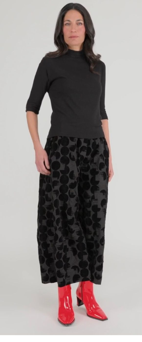Woman in black top, floral pants, and red boots, standing against a white backdrop.