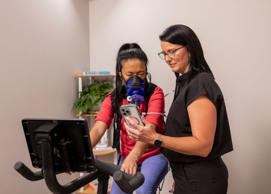 A woman is standing next to a woman on an exercise bike.