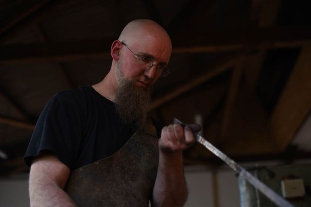 Portrait von Sören Binger bei der Arbeit. Das geschmiedete Stück Stahl wird von ihm in Augenschein genommen und geprüft.