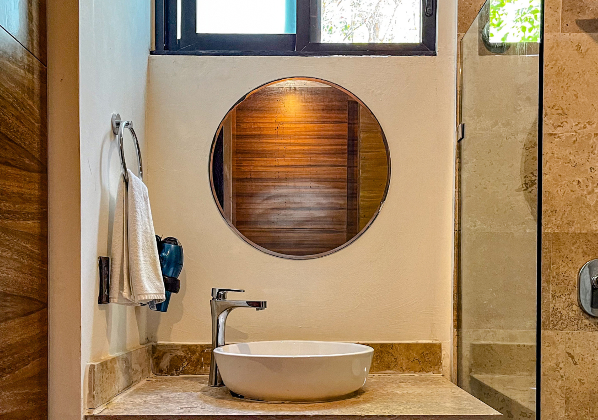 Mueble de baño con espejo redondo, lavabo sobre encimera y grifería cromada. Paredes y encimera en tonos tostados.