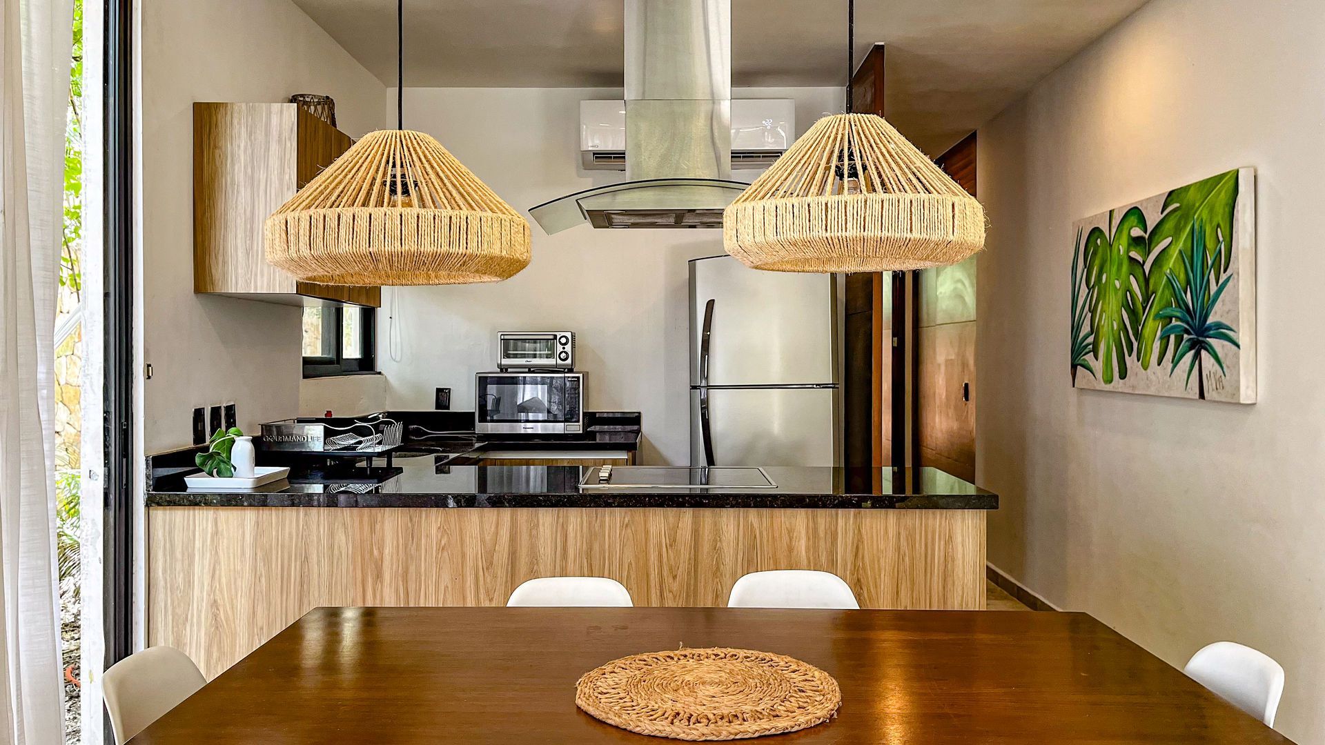 Cocina con mesa de comedor, gabinetes de madera, electrodomésticos de acero inoxidable y dos lámparas colgantes tejidas.