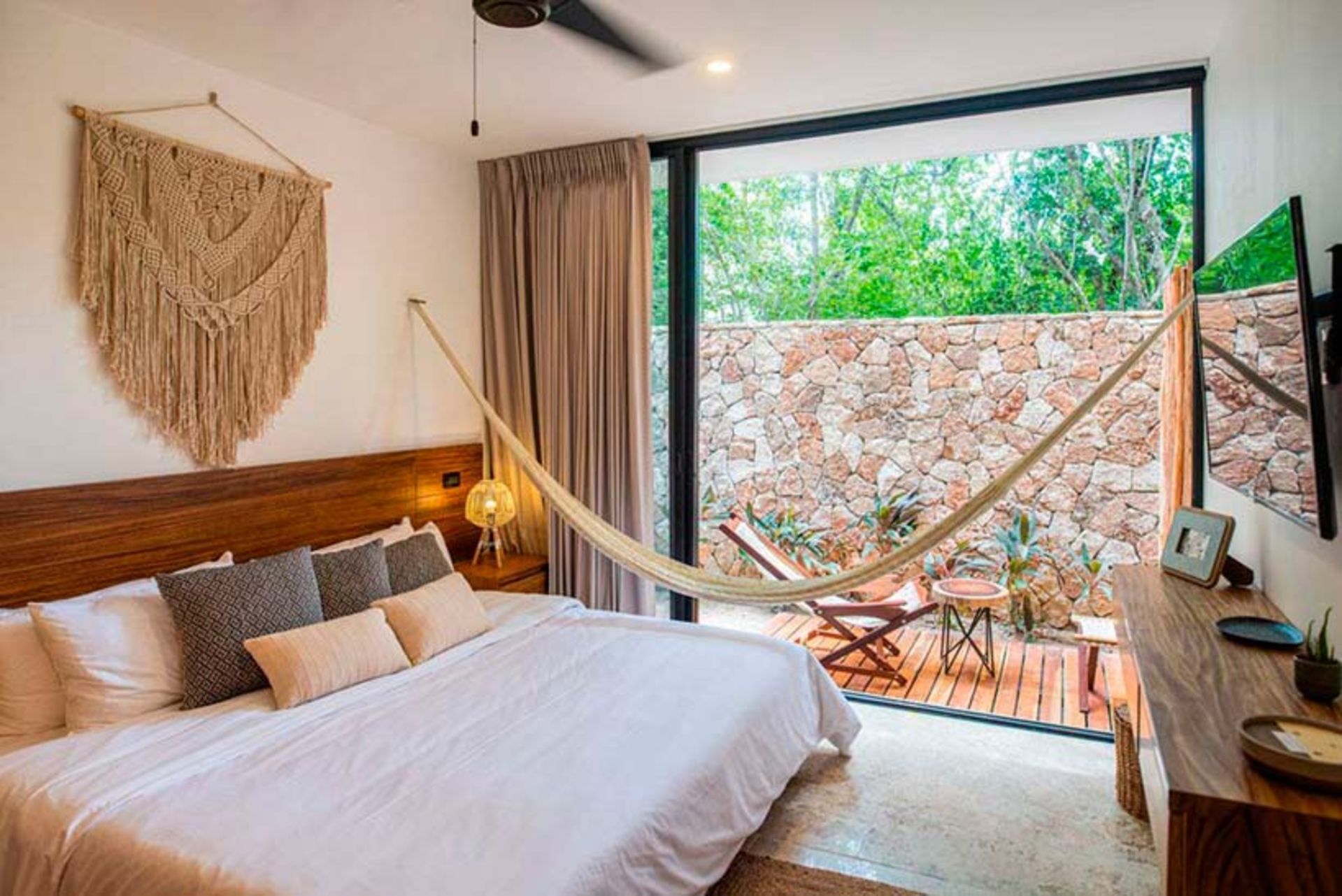 Dormitorio con cama, hamaca, puerta abierta al patio, pared de piedra y tapiz de macramé.