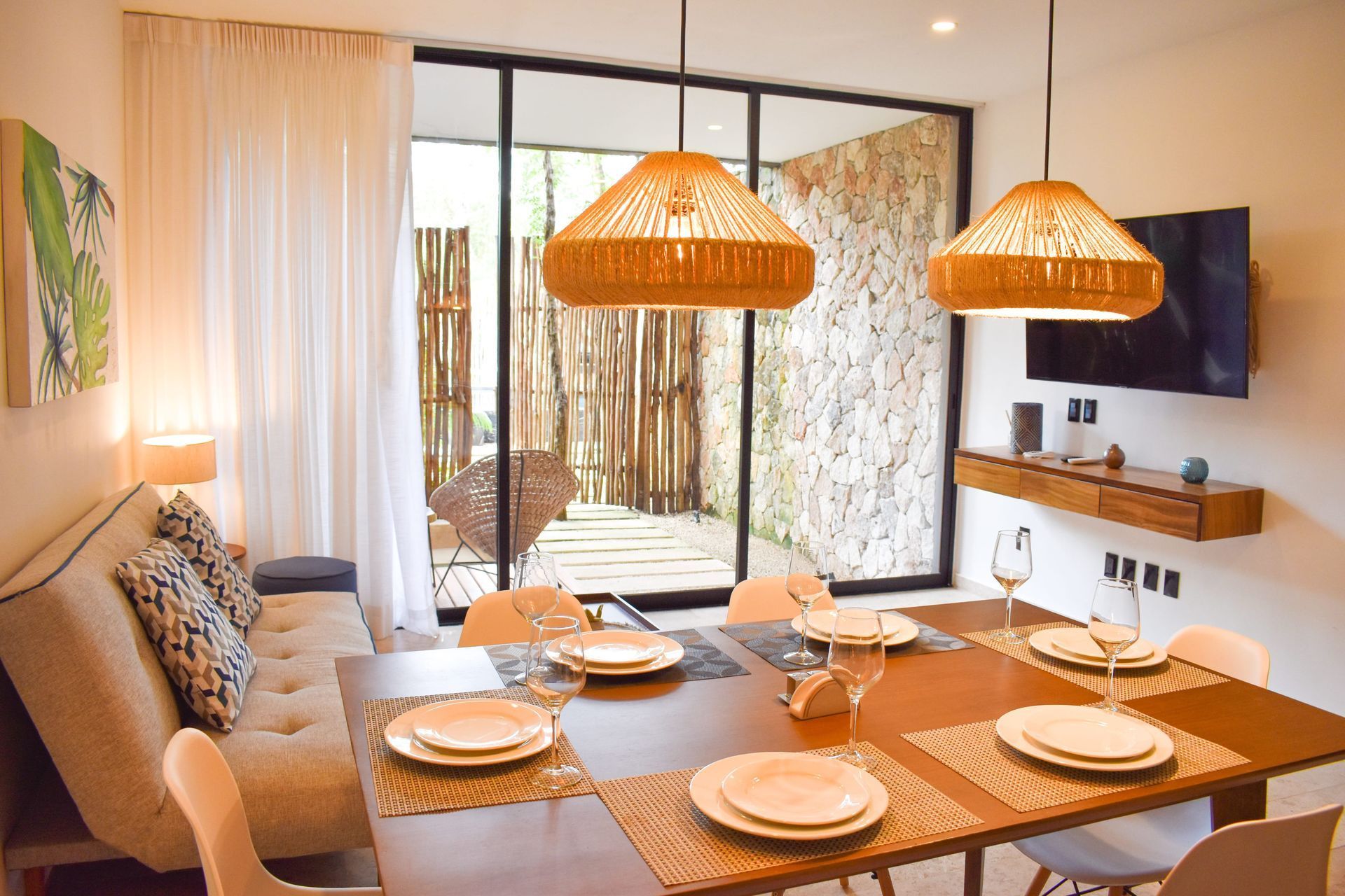 Comedor con mesa puesta para seis, un sofá largo y una puerta de vidrio que conduce a un patio al aire libre con vegetación.