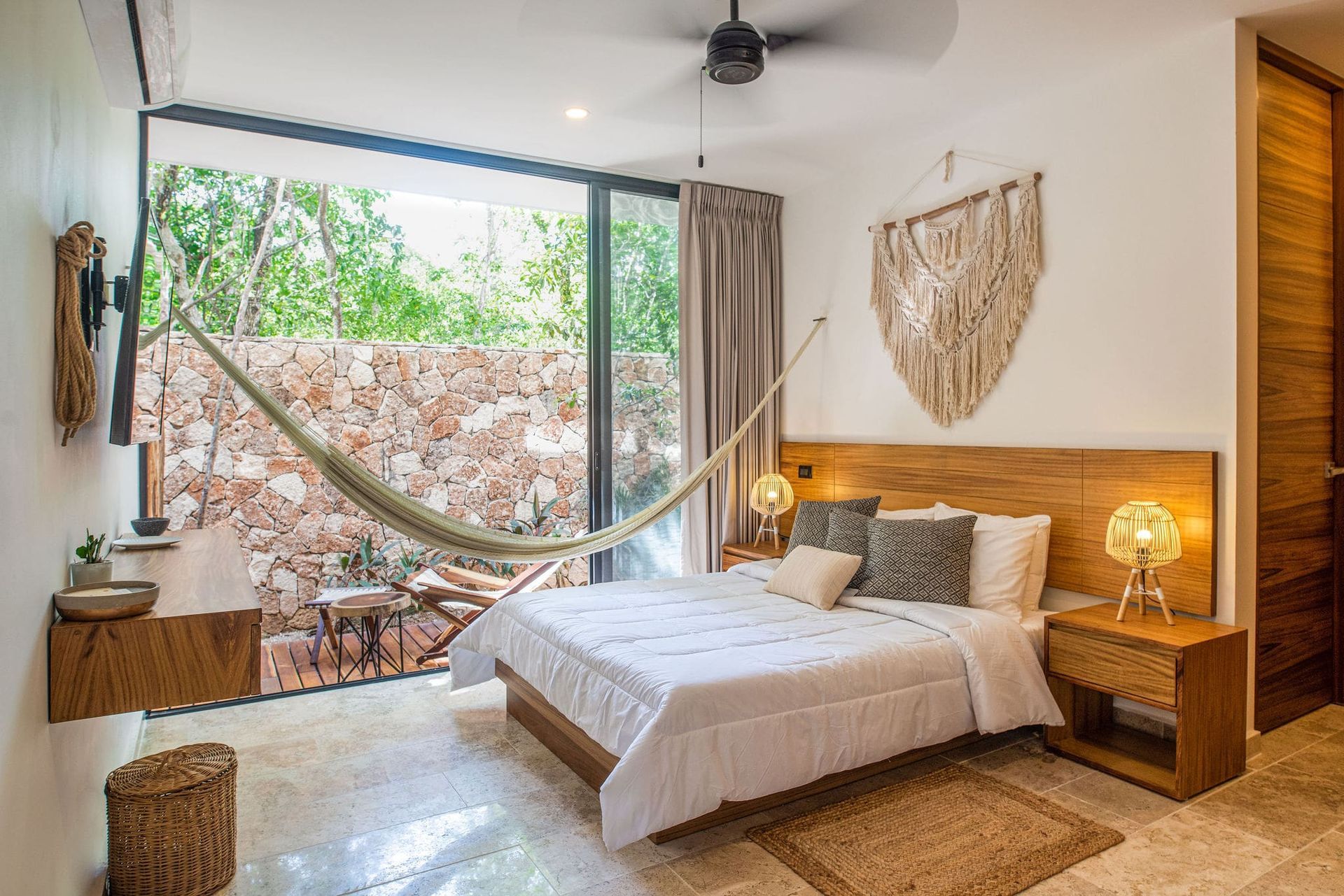 Dormitorio con hamaca, cama y ventana abierta con vista a un muro de piedra y exuberante vegetación.