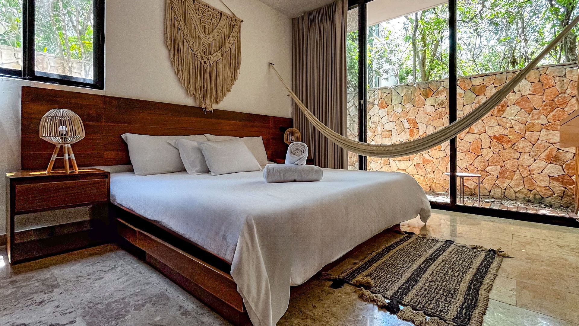 Dormitorio con cama de madera, pared de piedra natural, hamaca y gran ventanal con vista a los árboles.