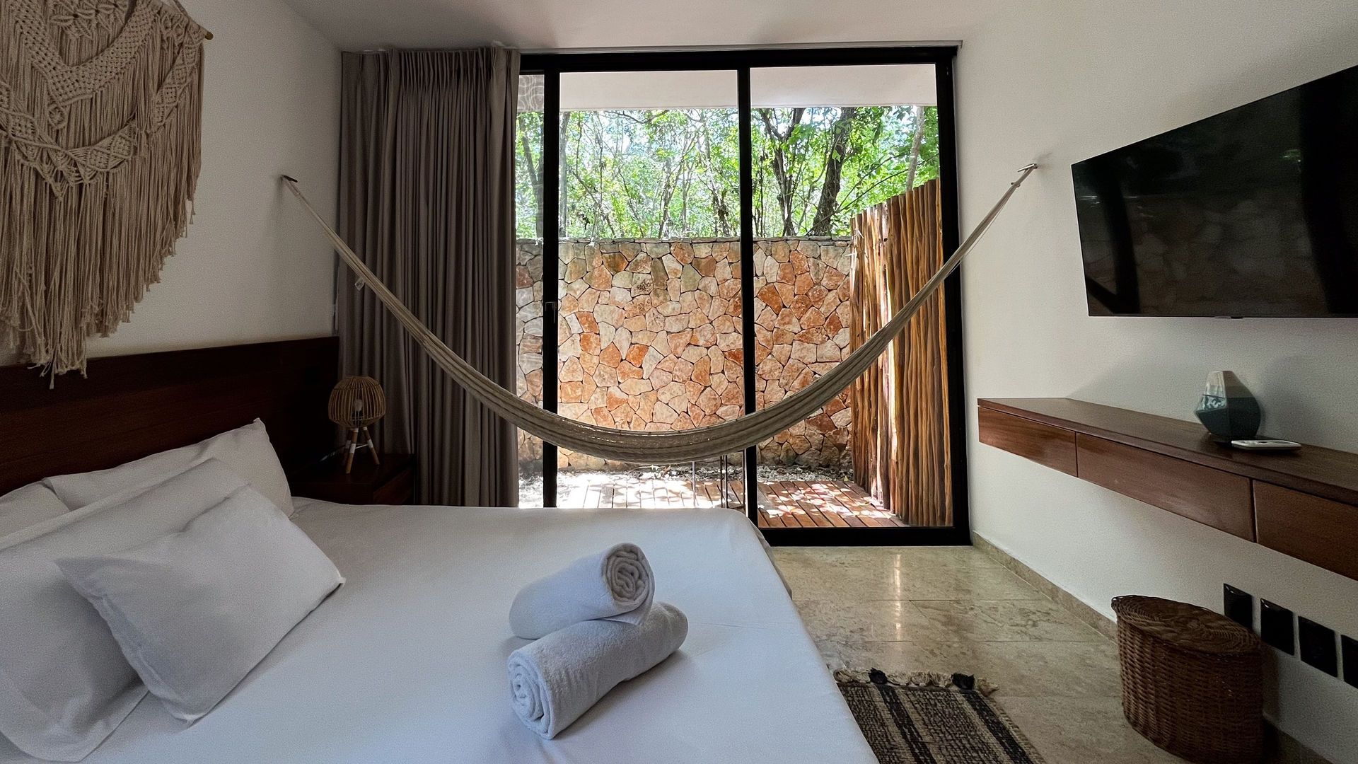 Dormitorio con cama, hamaca y puerta corrediza a un patio de piedra. Hay un televisor colgado en la pared.