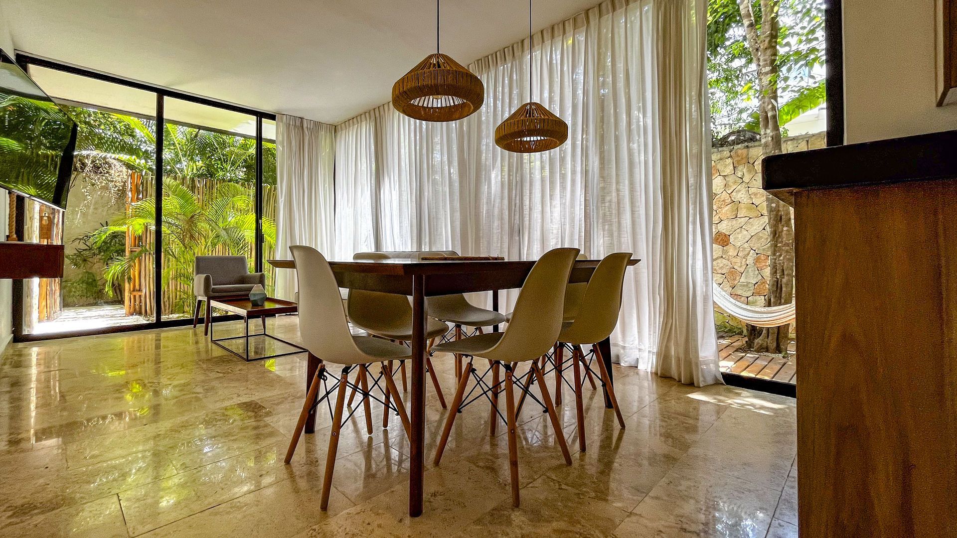 Comedor con mesa, sillas, amplios ventanales y cortinas blancas transparentes, con vista al verde exterior.