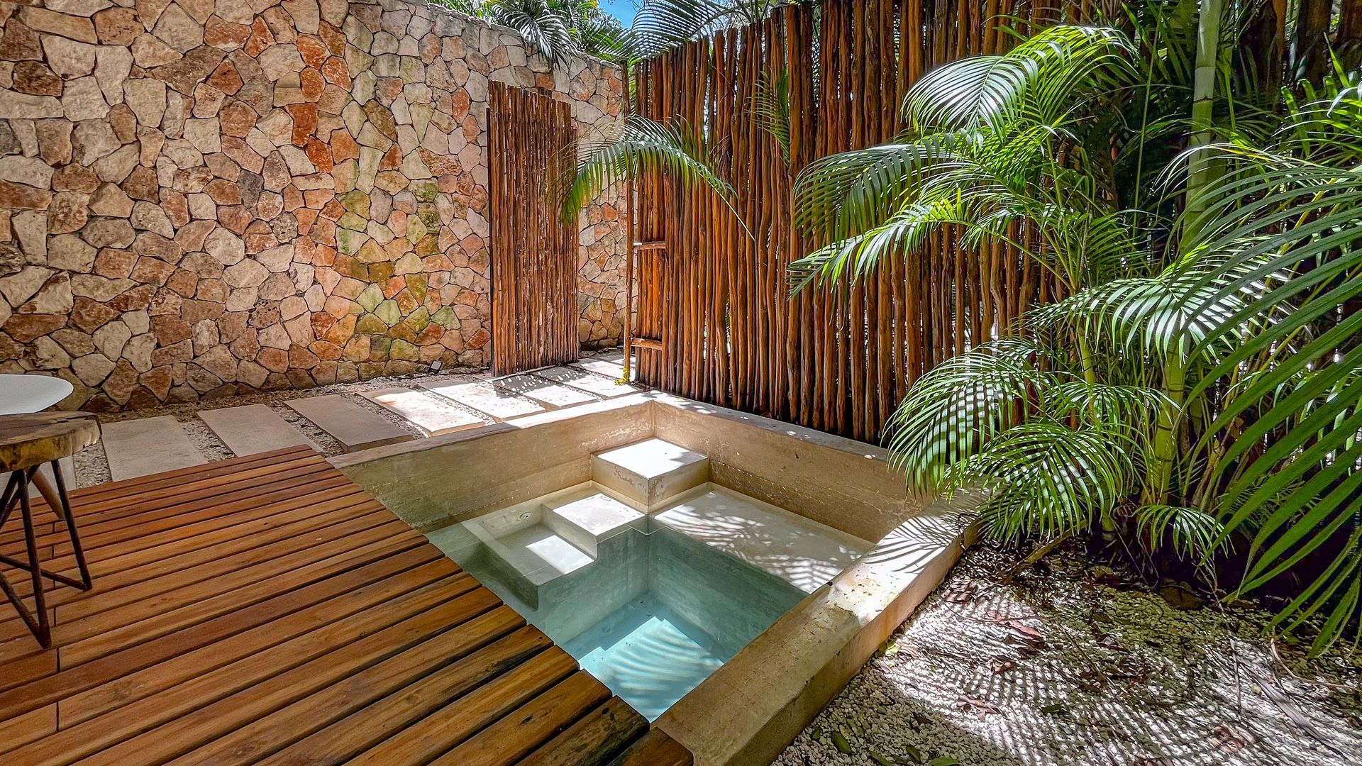 Pequeña piscina al aire libre rodeada de un muro de piedra, una valla de madera y exuberantes plantas tropicales.