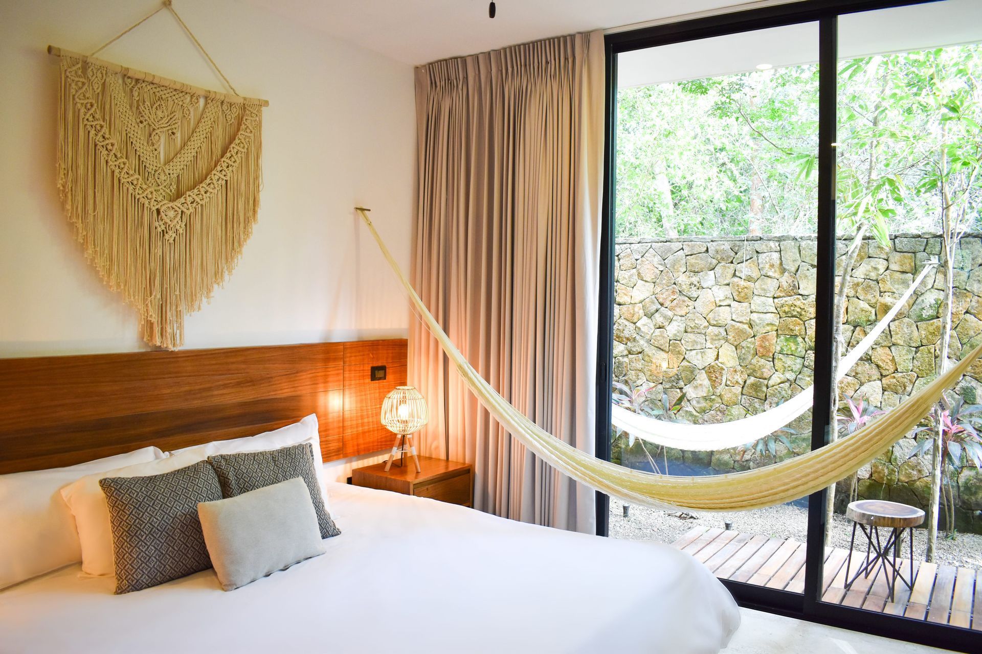 Dormitorio con cama, tapiz de macramé, hamaca y puerta corrediza de vidrio con vista a la vegetación.