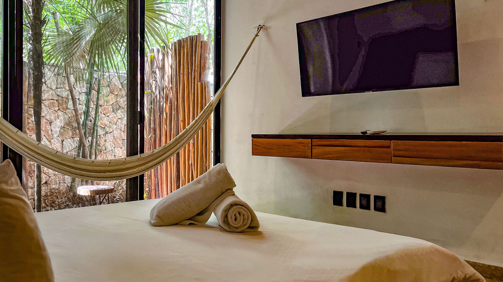 Habitación con cama, hamaca, televisor y vistas a la vegetación. Paredes blancas y detalles en madera.