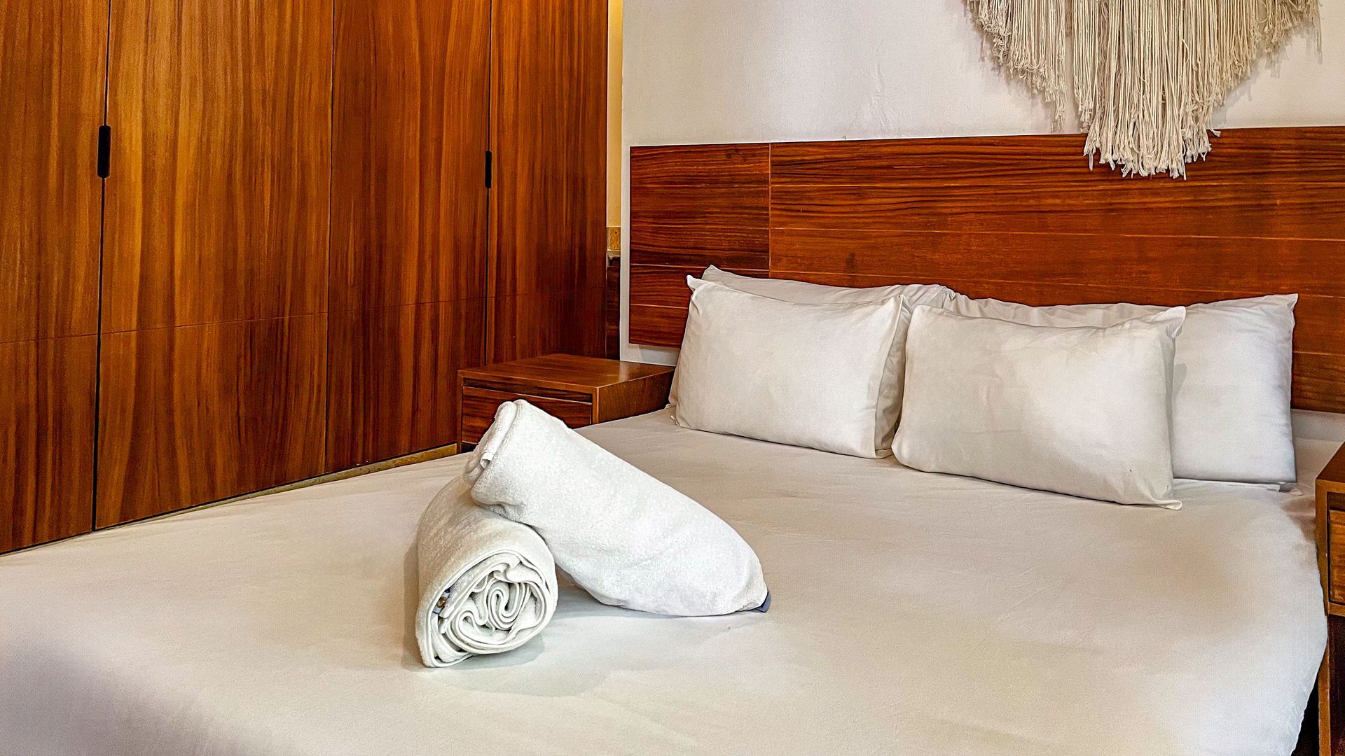 Dormitorio con cabecero de madera, ropa de cama blanca, toallas enrolladas y armario de madera.
